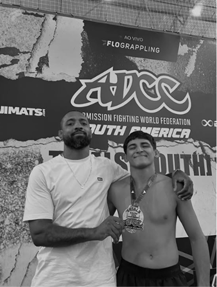 Professor e aluno segurando uma medalha em frente ao painel do ADCC South American Championship.